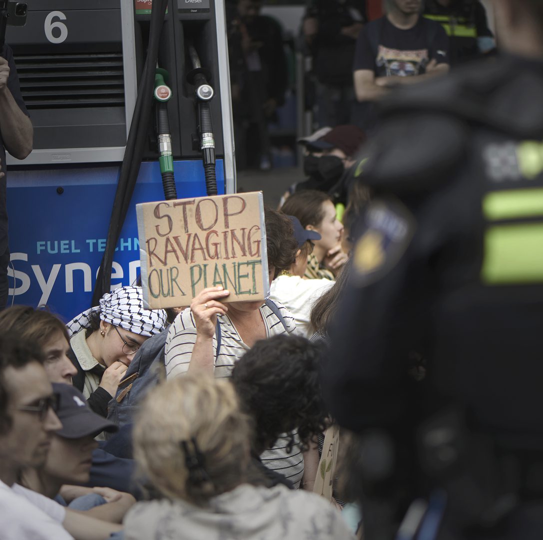 Zittende demonstranten bij Esso-tankstation tijdens klimaatprotest Den Haag met bord ‘Stop ravaging our planet’
